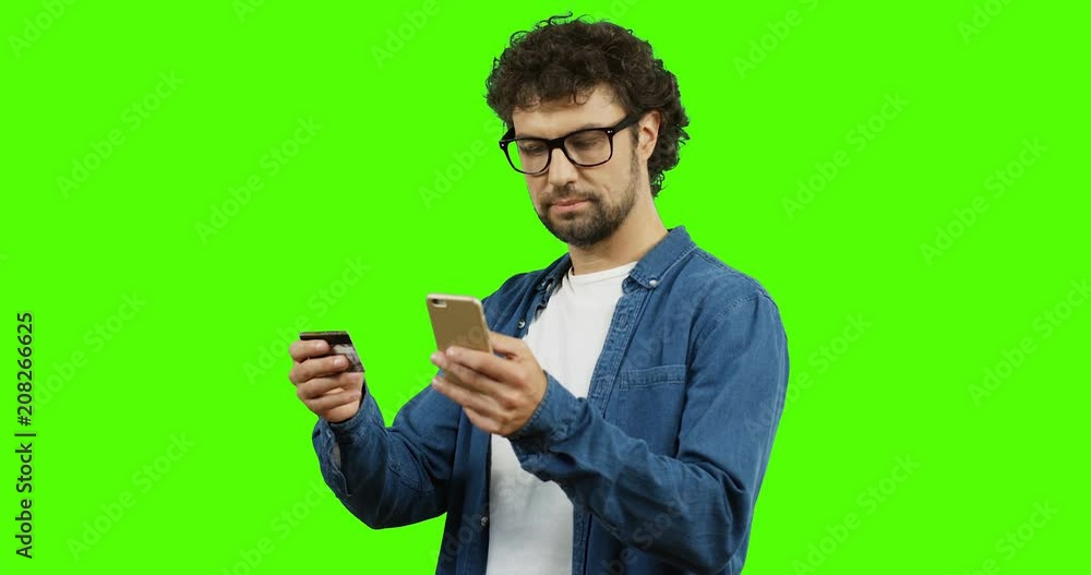 Young Caucasian man in glasses typing credit card number on the smartphone while shopping online. Chroma key. Green screen. Young Caucasian man in glasses typing credit card number on the smartphone while shopping online. Chroma key. Green screen.