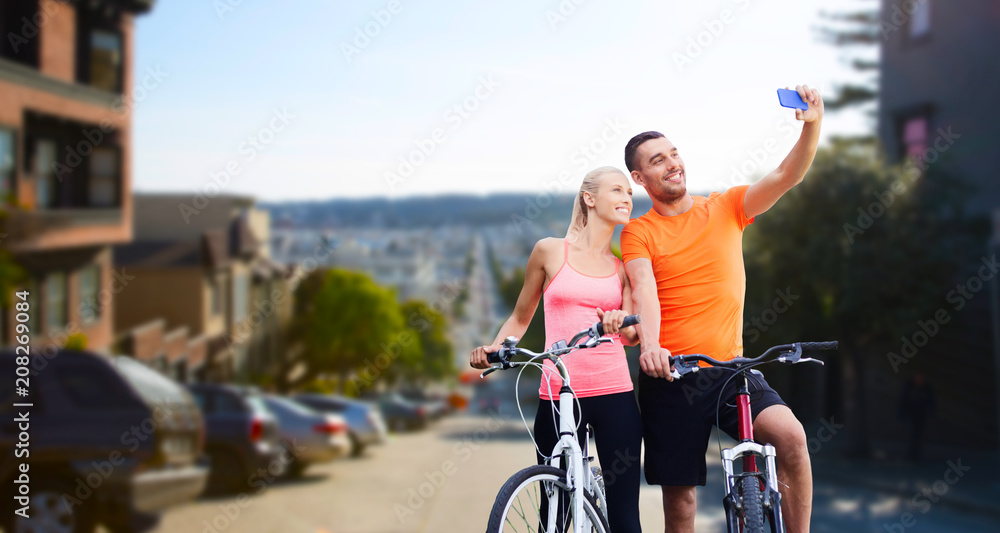 Obraz premium fitness, sport, technology and healthy lifestyle concept - happy couple riding bicycles and taking selfie by smartphone over san francisco city background