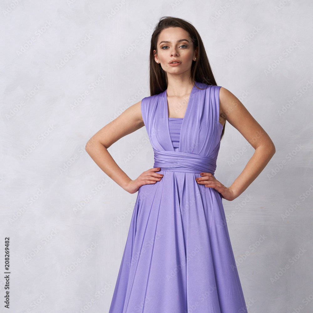 Beautiful long haired young woman dressed in stylish purple bandeau maxi dress posing against white wall on background. Elegant brunette female model demonstrating evening outfit in studio.