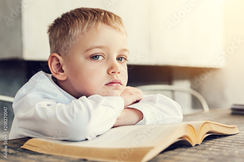 Little pupil. Pretty little boy looking tired and sad while sitting at the table with a big book