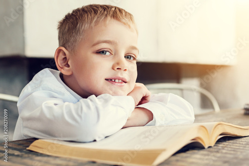 Cheerful boy. Pleasant smiling friendly boy sitting at the table with his arms on the book