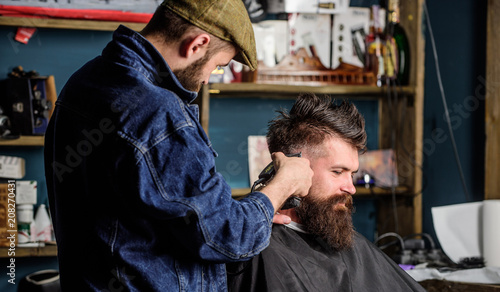 Hipster lifestyle concept. Hipster client getting haircut. Barber with hair clipper works on hairstyle for bearded man barbershop background. Barber with clipper trimming hair on temple of client