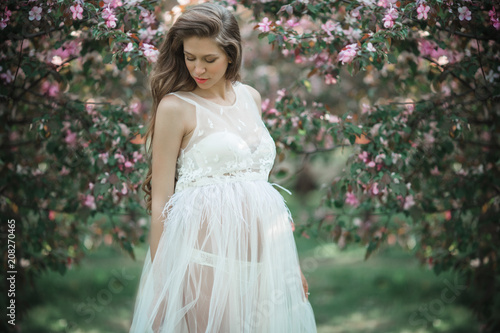 Pregnant pretty woman is wearing white fashion dress posing in pink blossom apple garden, summer time