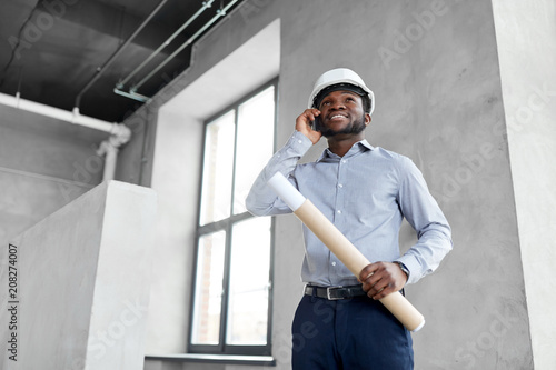 architecture, construction business and people concept - african american architect in helmet with blueprint calling on smartphone