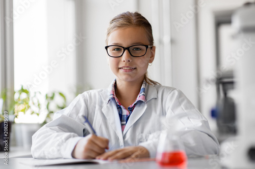 education, science and children concept - girl studying chemistry at school laboratory and writing to workbook