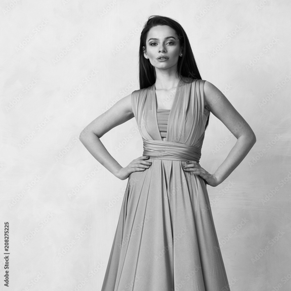 Beautiful long haired young woman dressed in stylish purple bandeau maxi dress posing against white wall on background. Elegant brunette female model demonstrating evening outfit in studio. Beautiful long haired young woman dressed in stylish purple bandeau maxi dress posing against white wall on background. Elegant brunette female model demonstrating evening outfit in studio.