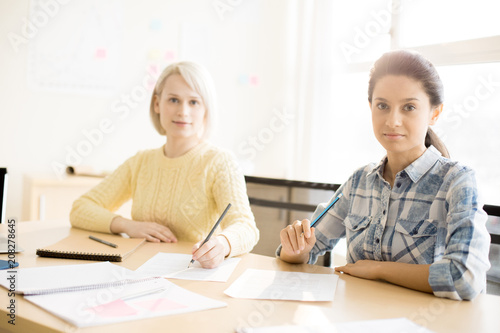 Young females sitting at desk with notebook and papers making notes in office and looking at camera