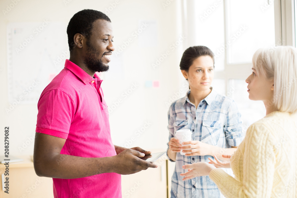 Obraz premium Side view of two young women and smiling black man in bright pink t-shirt conversing with interest standing in office