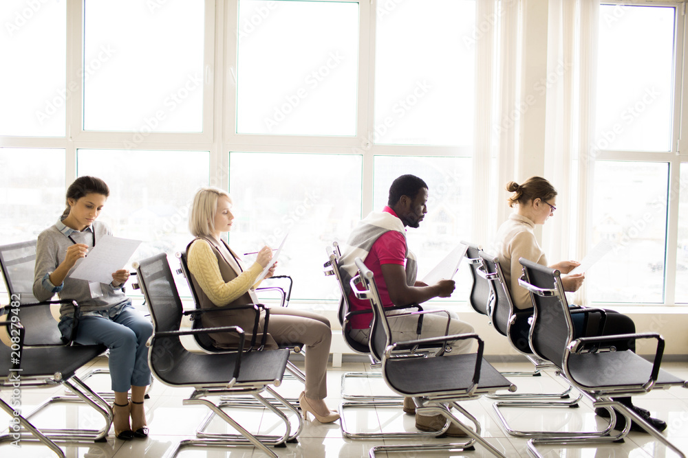 Side view of three young women and black man doing business tasks with concentration sitting one on each row in light conference hall Side view of three young women and black man doing business tasks with concentration sitting one on each row in light conference hall