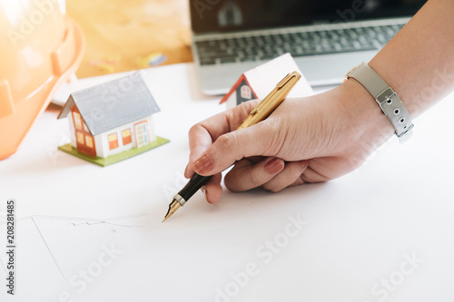 Close up of Young Female Architect Freehand Drawing, Civil engineer sketching a blueprint of construction project with small house model and safety helmet in office, Construction Concept.