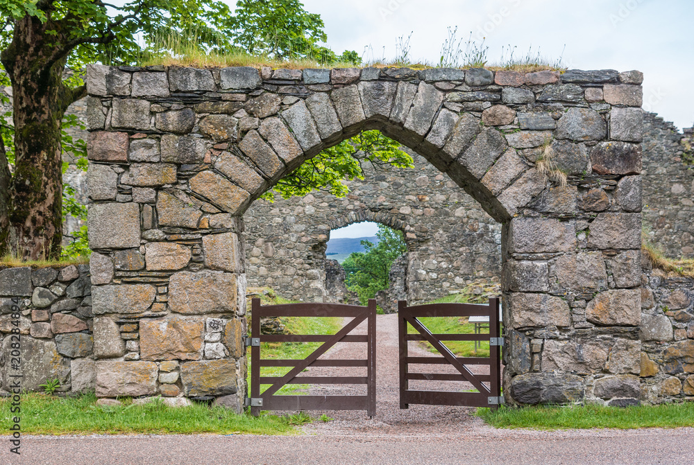 Naklejka premium Torlundy, Scotland - June 11, 2012: Natural Stone gate with triangular bow to the domain of Inverlochy Castle near Fort William. Green grass and tree foliage. Gray sky.