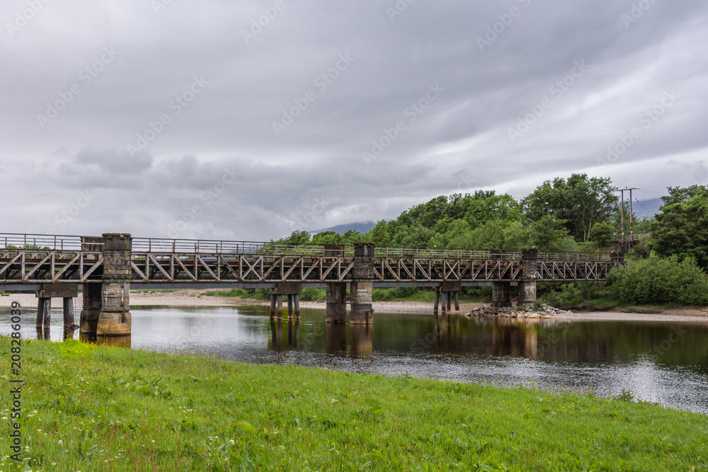 Torlundy, Scotland - June 11, 2012: Railway bridge over the gray River Lochy behind Inverlochy Castle under heavy dark skies. Grassland on rocky shore. River protects the castle at three sides.