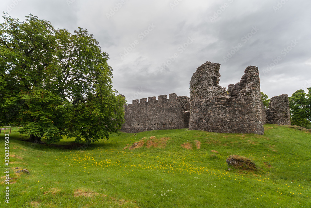 Torlundy, Scotland - June 11, 2012: Ruins of Natural Stone ramparts With Comyn Tower of Inverlochy Castle near Fort William. Outside view with tree, green grass and light gray sky.
