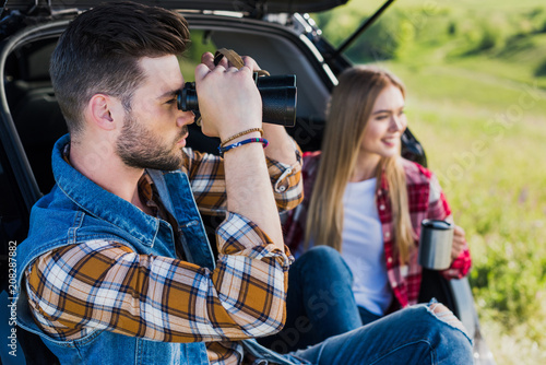 side view of male tourist looking through binoculars while his smiling girlfriend sitting near with coffee cup on car trunk