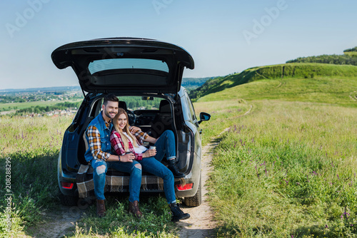 happy couple of stylish tourists with coffee cups sitting on car trunk in rural field