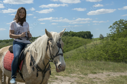Photo of the small woman on a big horse in summer field. Image of happy female sitting on purebred horse and looking at camera outdoors