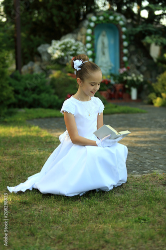 Śliczna dziewczynka w białej sukience modli się z książeczki do nabożeństwa na tle kapliczki.