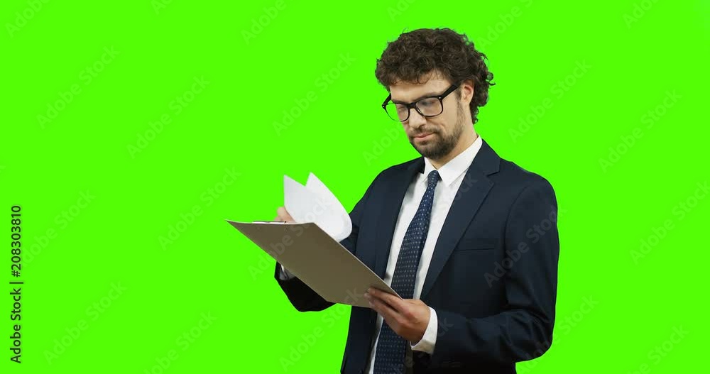 Handsome Caucasian man in the glasses, suit and tie looking at the documents in his hands and doing yes gesture with his hand to the camera on the green screen. Chroma key. Handsome Caucasian man in the glasses, suit and tie looking at the documents in his hands and doing yes gesture with his hand to the camera on the green screen. Chroma key.