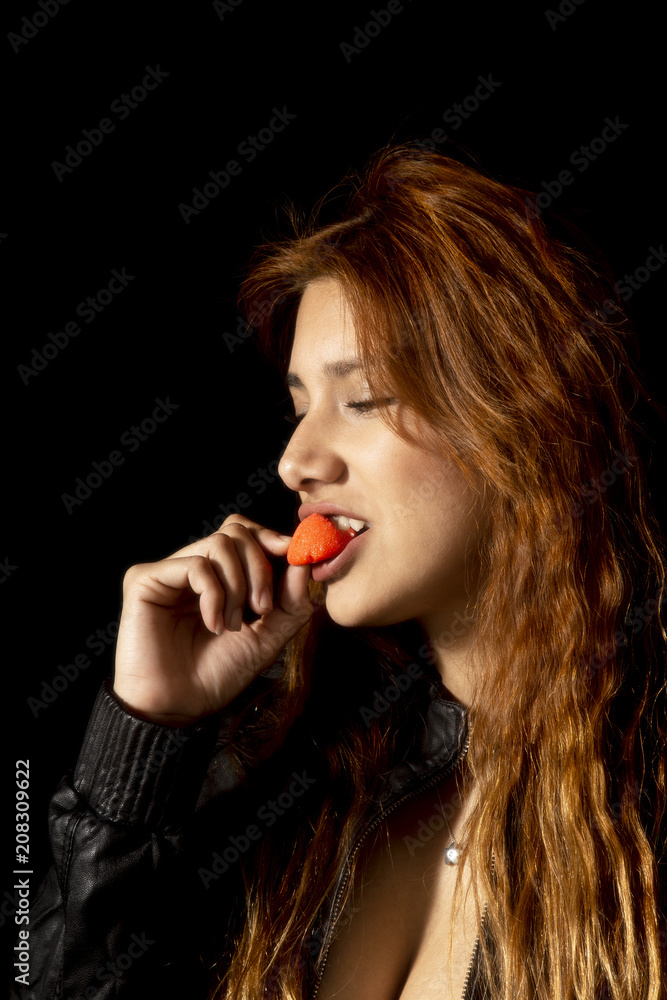 Redhead latin girl with black jacket on black background eating a strawberry Redhead latin girl with black jacket on black background eating a strawberry