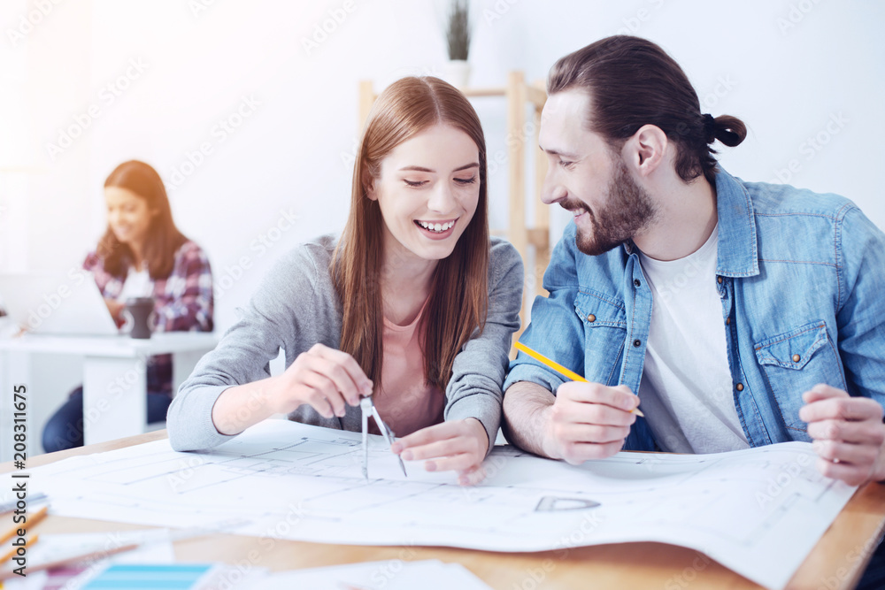 Obraz premium Work with pleasure. Charming girl keeping smile on her face and using pair of compasses while listening her partner