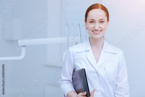 Visit my clinic. Delighted intern keeping smile on her face and holding folder while feeling happiness