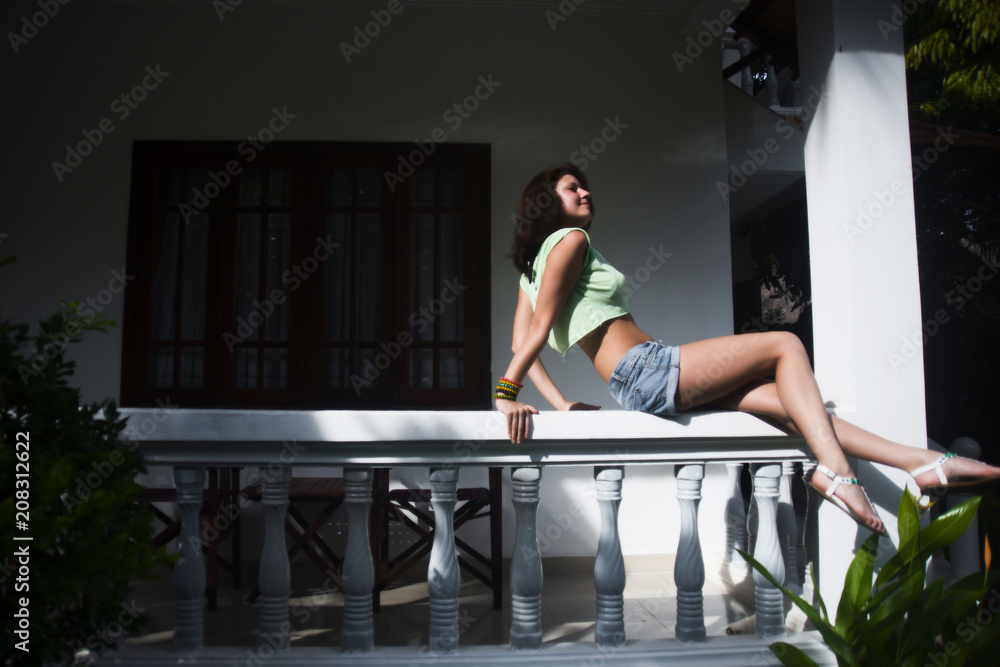 Obraz premium Beautiful girl in shorts and a t-shirt sexy sitting on a white railing on a hot summer day in the background of the cottage