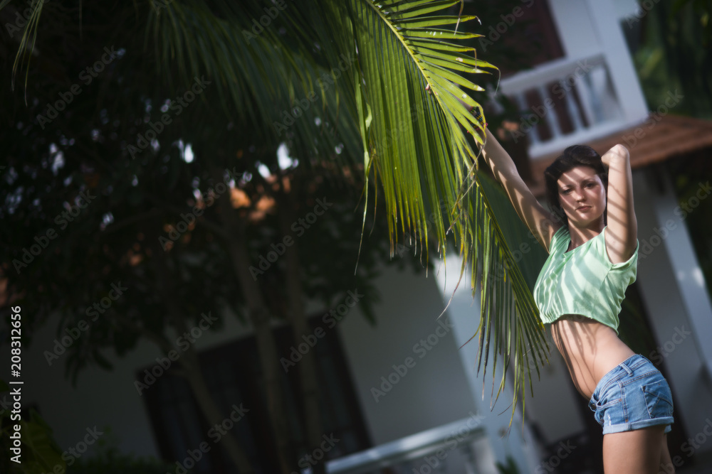 Young beautiful girl in shorts posing with palm leaf in the summer against the cottage