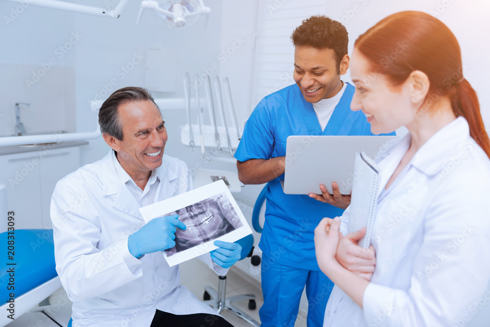 Funny moment. Professional dentist keeping smile on his face and sitting in his cabinet while holding picture in both hands Funny moment. Professional dentist keeping smile on his face and sitting in his cabinet while holding picture in both hands