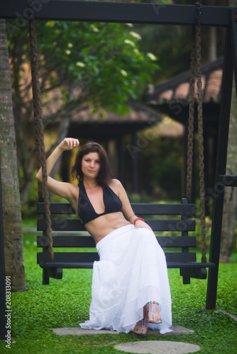 Beautiful woman in white dress sitting on a swing on a green lawn in summer at the resort © Vladimir