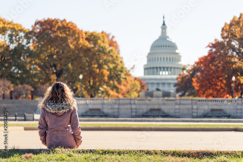 Young woman in coat sitting looking at view of United States Congress Capitol building, golden orange yellow foliage autumn fall trees on street during sunny day in Washington DC photo