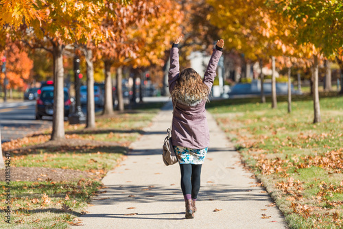 Young happy woman, arms raised on sidewalk street walking in Washington DC, USA in alley of golden orange yellow foliage autumn fall trees on road during sunny day photo