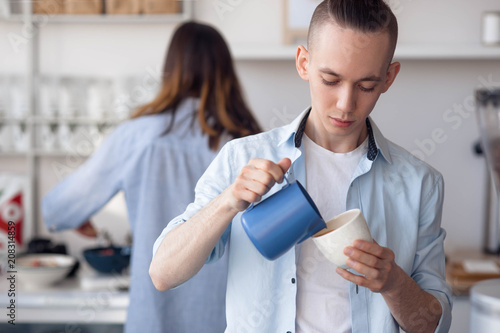 The men with stylish hairstyle stands and cooks cappuchino at work. He pours milk in coffee