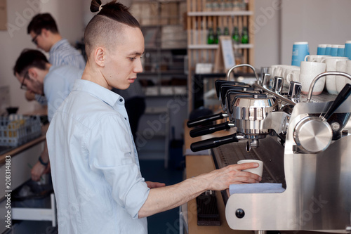 A young guy works as a Barista  he stands at the coffee machine and fills a Cup with hot coffee