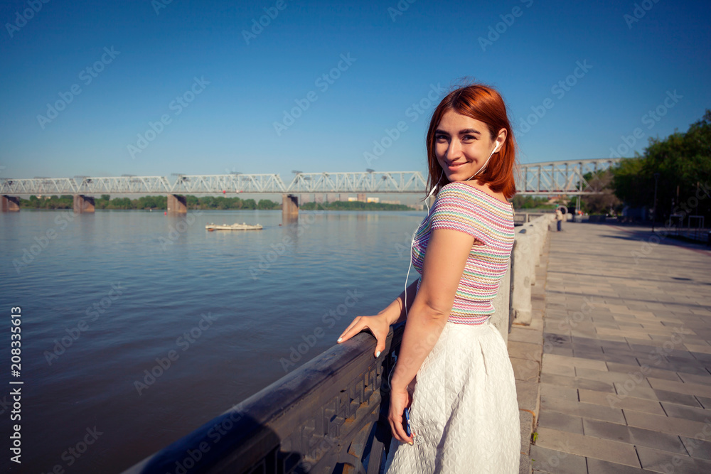 A young red-haired woman in a white romantic skirt, a pink top listens to music on the phone and looks at the river on the city waterfront on a summer day