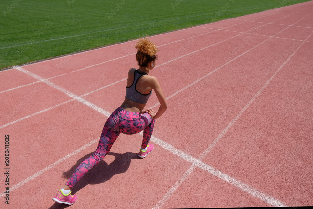 Exercises on the sports field. A young dark-skinned girl in a gray T-shirt, pink pants and sneakers holds her hands on one knee on the starting line of the stadium's racetrack. View from the back.