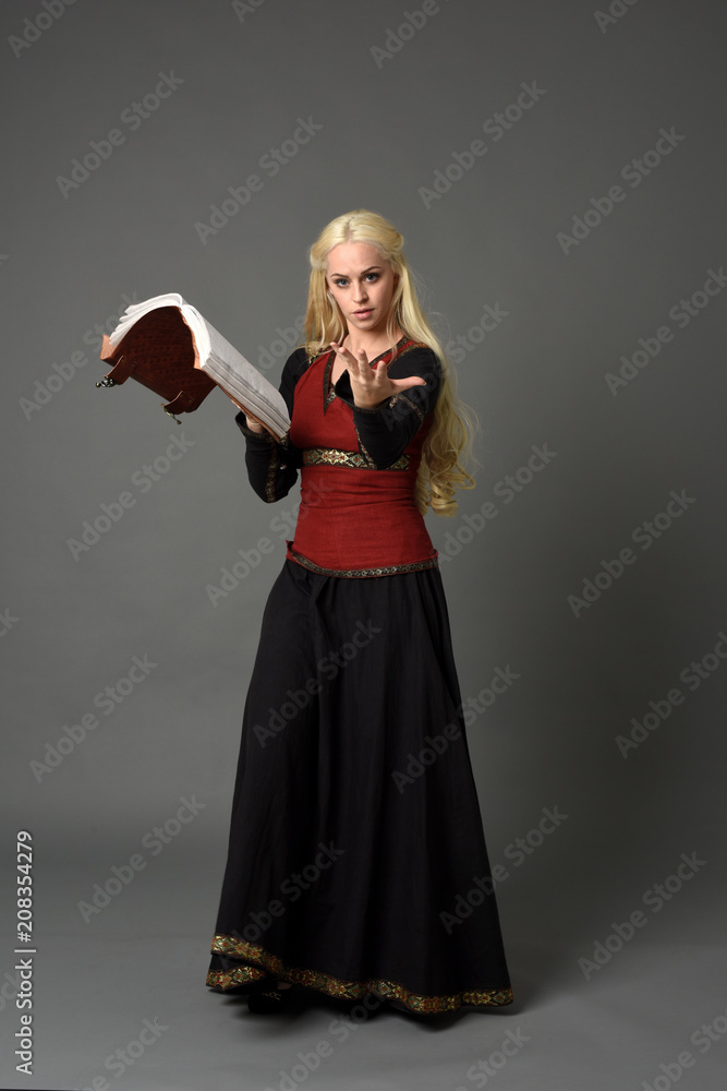 full length portrait of pretty blonde lady wearing a red and black fantasy medieval gown, holding a book. standing pose on grey background. full length portrait of pretty blonde lady wearing a red and black fantasy medieval gown, holding a book. standing pose on grey background.