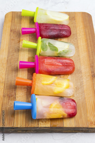 Homemade colorful fruity popsicle on a wooden board and against a white stone background. Strawberry, Lemon, Lemon with mint, Orange, Cherry, Multifruit