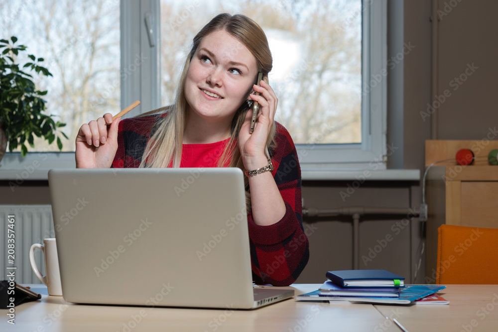 Obraz premium A smiling blonde woman sitting at a laptop and talking over a cell phone.