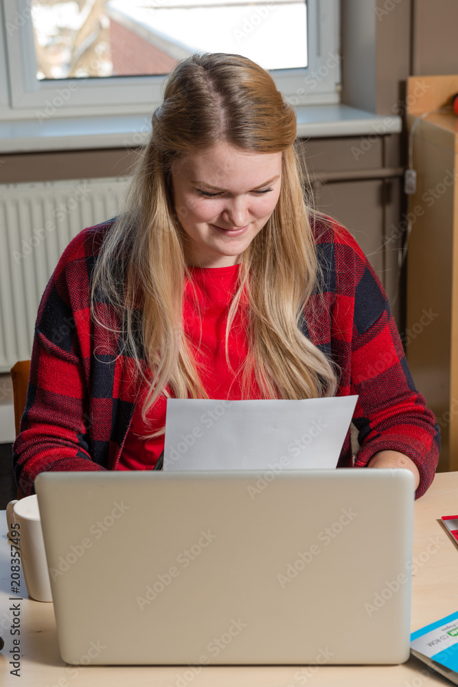 Fototapeta premium Smiling blonde woman sitting at a laptop drinking tea and working.