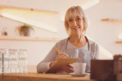 Organized waiter. Pleasant senior woman in an apron checking customers orders in her notes and smiling at the camera while working as a waiter in a cafe