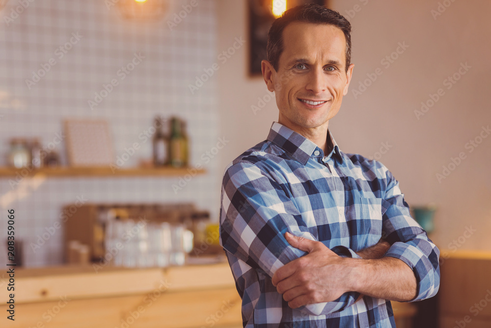 Obraz premium Upbeat owner. Cheerful young male cafe owner in a checked shirt folding his arms across his chest and smiling while posing for the camera in his own cafe