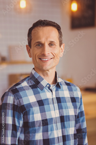 Pleased customer. Pleasant upbeat young man in a checked shirt posing for the camera and smiling while standing near the bar counter in the cafe