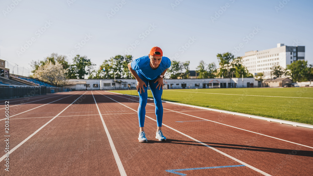 Fototapeta premium Athletic handsome male relaxing after intense workout and running on stadium. Athlete taking rest with hands resting on knees after a jogging and run standing on race track outdoor. People and sport
