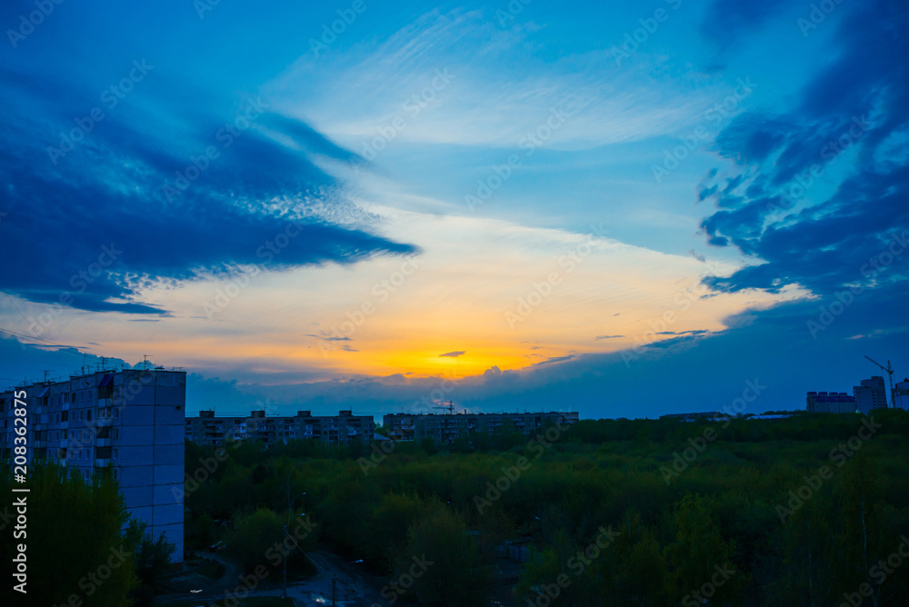 Atmospheric blue cloudy sky behind silhouettes of city buildings. Cobalt and orange background of sunrise with dense clouds and bright yellow sunny light for copy space. Cyan heaven above clouds. Atmospheric blue cloudy sky behind silhouettes of city buildings. Cobalt and orange background of sunrise with dense clouds and bright yellow sunny light for copy space. Cyan heaven above clouds.