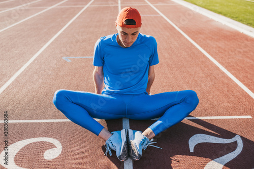 Sportive Caucasian athlete doing stretching exercises sitting on the running track. Runner male sitting on running track with feet joined together preparing for a run or relaxing after workout.