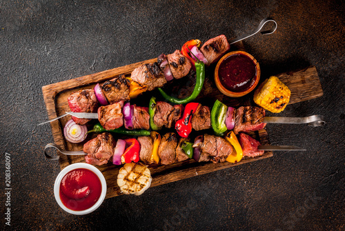 Fresh, home-cooked on the grill fire meat beef shish kebab with vegetables and spices, with barbecue sauce and ketchup, on a dark background on a wooden cutting board above copy space