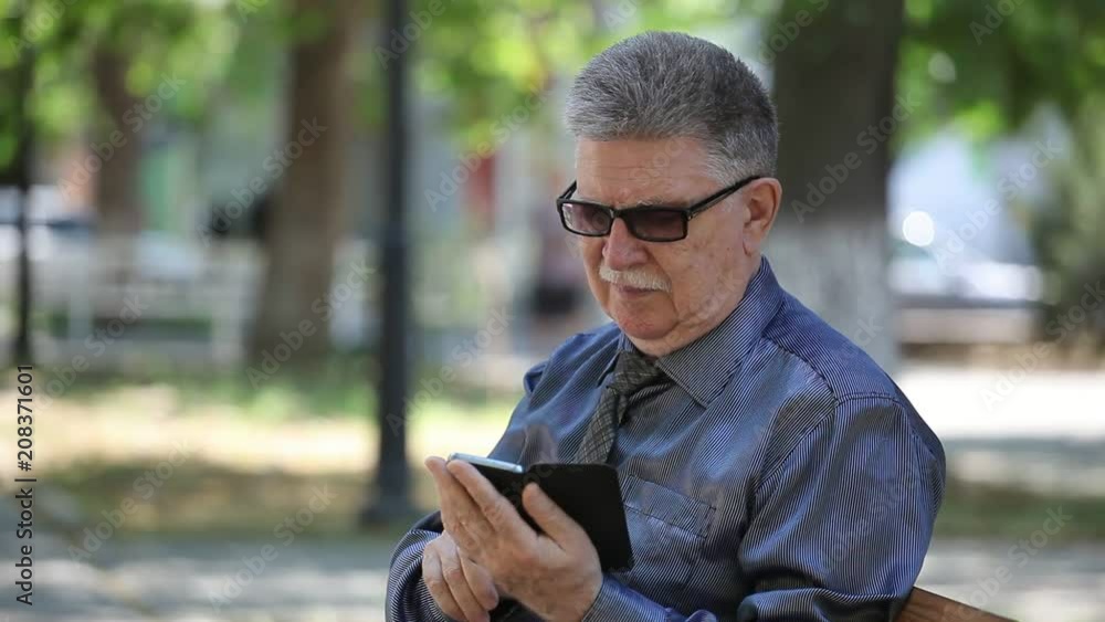 Portrait of a grey-haired man with mustashe in a necktie surfing internet on his phone sitting in a splendid alley on a sunny day in summer Portrait of a grey-haired man with mustashe in a necktie surfing internet on his phone sitting in a splendid alley on a sunny day in summer
