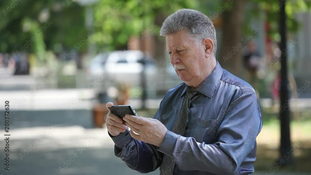 Portrait of a brave white-haired man with mustashe in a necktie seeking info on his smartphone in a green street on a sunny day in summer Portrait of a brave white-haired man with mustashe in a necktie seeking info on his smartphone in a green street on a sunny day in summer