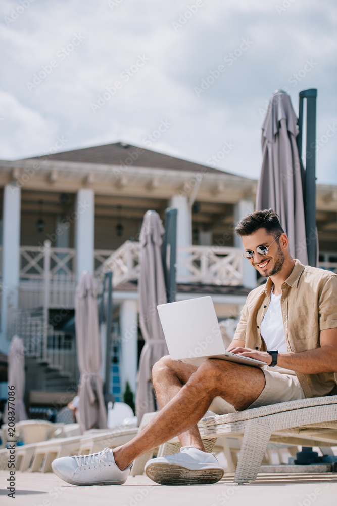 smiling young man in sunglasses sitting on chaise longue and using laptop