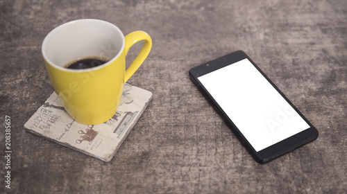 Black phone on a wooden table next to yellow cup of coffee mock up